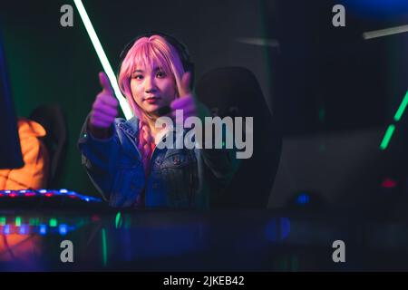 Teenage asian pink-haired girl sitting in gaming chair by a desk giving thumbs-up to the camera. Gaming computer setup. Colored lights. High quality photo Stock Photo