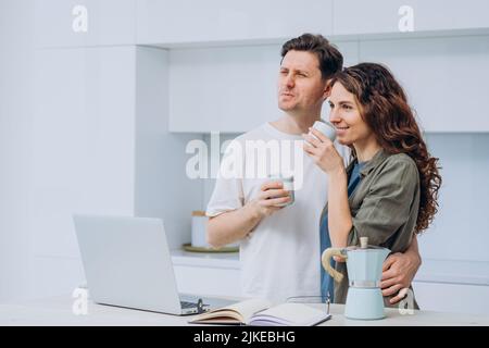 Married couple of freelancers took a coffee break in a bright kitchen standing at a table with a laptop. Happy man and woman stand embracing and drinking aromatic coffee. Stock Photo