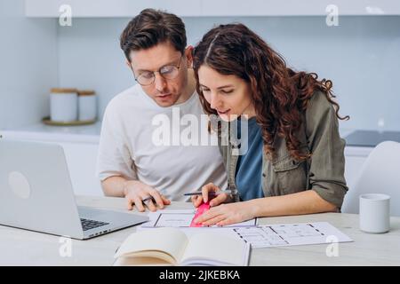 Couple sits in kitchen drinks coffee for breakfast and talks about drawings of apartment renovation making notes in laptop and notebook Stock Photo