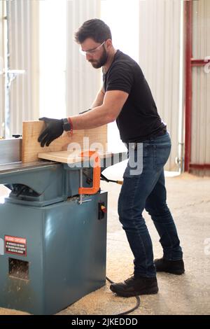 hands of a young carpenter working on an Electric Benchtop Jointer machine cutting a wooden board in a carpentry workshop using safety black gloves Stock Photo