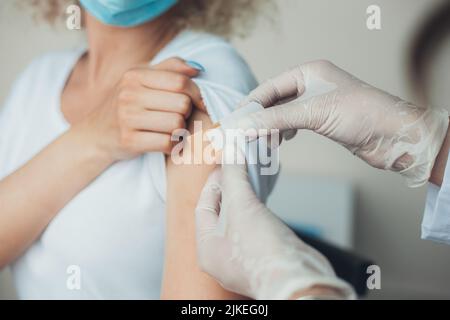 Doctor in protective gloves putting an adhesive bandage on young woman's arm after injection of vaccine. Vaccination against coronavirus while COVID Stock Photo