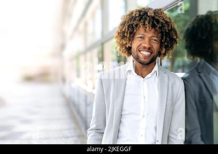 Happy handsome confident positive african american or brazilian man with curly hair, dressed in stylish formal wear, standing outdoors, looking at the camera, smiling friendly. Copy-space Stock Photo