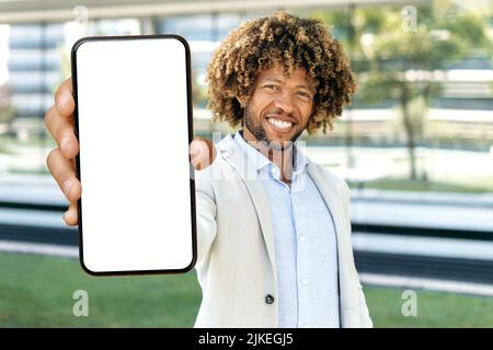 Positive attractive african american or brazilian curly haired man, stands outdoors, shows smart phone with blank white mockup screen with space for advertising, presentation, looks at camera, smiles Stock Photo