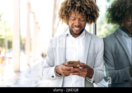 Busy friendly successful brazilian or african american curly haired man, stylishly elegantly dressed, stands outdoors near the business center, using mobile phone, messaging with friends, smiling Stock Photo