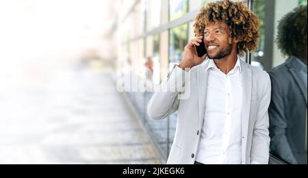 Positive busy african american or brazilian curly haired man, wearing stylish business clothes, stands outdoors near business center, having conversation by smartphone, looking away, smiling friendly Stock Photo