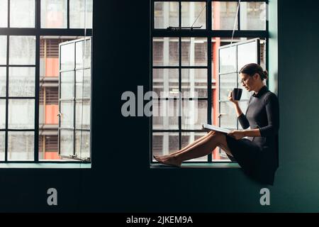 Asian woman sitting on window sill reading a book and drinking coffee. Female relaxing indoors with magazine and having coffee. Stock Photo
