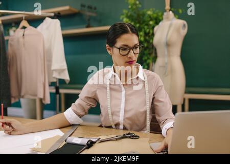 Female dressmaker drawing dress patterns with laptop. Creative fashion designer using laptop in her studio. Stock Photo