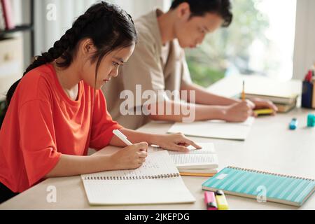 Concentrated schoolgirl reading novel and writing synopsis in copybook at English class Stock Photo