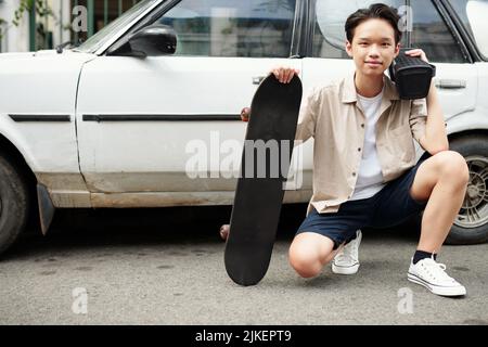 Positive teenage boy spending time outdoors with kating and listening to music through speaker Stock Photo