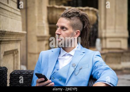 Young successful hipster caucasian man with dreadlocks in a blue suit with a smartphone in his hands sits in a street restaurant looks away. Stock Photo