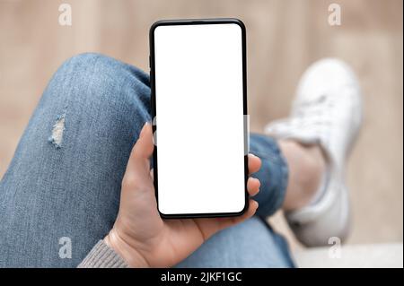 Top view mockup image of woman holding mobile phone with blank white screen. woman using cellphone with while sitting cross-legged on sofa at home. Mo Stock Photo