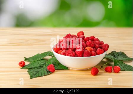 Plate with ripe red raspberries stands on wooden table against blurred background of greenery. Raspberry fruits in plate on wood desk, healthy pile of Stock Photo