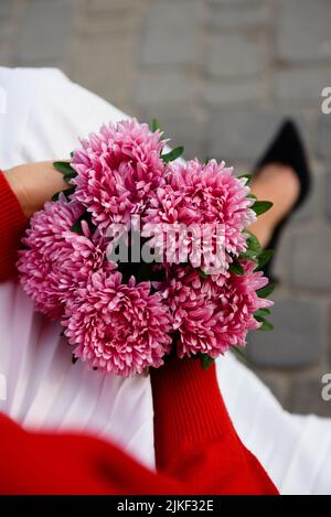Overhead view of a bouquet of dark pink asters in hands of a sitting woman in a white pleated skirt, red blouse and black high heels, selective focus. Stock Photo