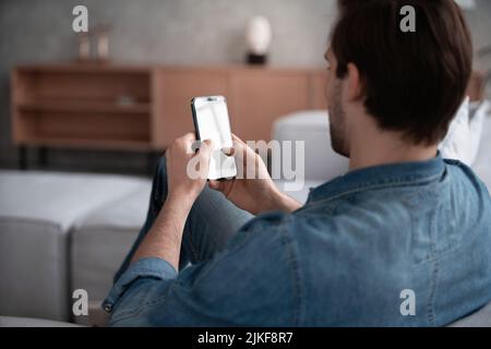 People And Technology Concept. Over The Shoulder View Of Unrecognizable Man Using Smart Phone With White Empty Screen For Mockup, Sitting At Home, Mak Stock Photo