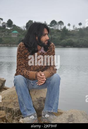 Full length of a young man with long hair and beard looking sideways while sitting on stone by the lake Stock Photo