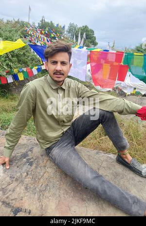 A handsome young guy looking at camera while sitting on rock with the background of Tibetan prayer flags in Rewalsar Stock Photo