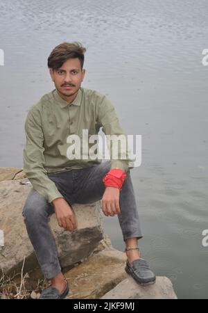 A young guy looking at camera while sitting on stone next to the lake Stock Photo