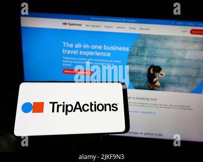 Person holding mobile phone with logo of American travel management company TripActions Inc. on screen in front of webpage. Focus on phone display. Stock Photo