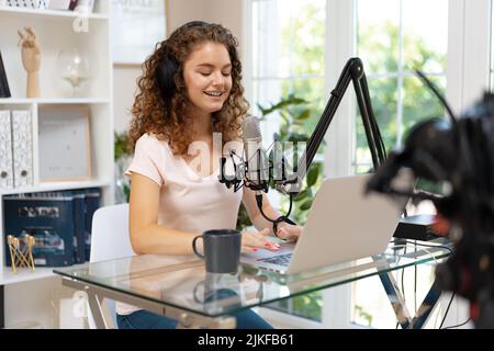Woman vlogger with headphones looking at laptop and recording video on professional camera Stock Photo
