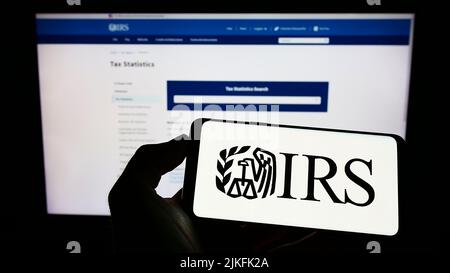 Person holding cellphone with logo of US Internal Revenue Service (IRS) on screen in front of webpage. Focus on phone display. Stock Photo