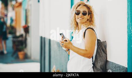 Happy female tourist pose for a picture using mobile phone connection against a white wall. Side portrait of woman smiling and writing on the cellular Stock Photo