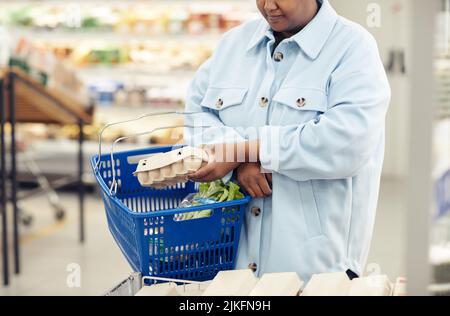Cropped shot of young black woman buying groceries in supermarket and putting eggs in shopping basket, copy space Stock Photo