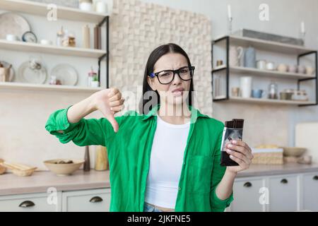 Woman at home disapproving of a bar of chocolate, looking at the camera and pointing a finger down, brunette at home in the kitchen in a green shirt and glasses Stock Photo