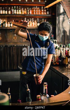 A female bartender with a mask pouring liquid into copper cocktail cup Stock Photo