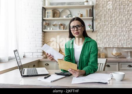 Portrait woman at home, received bad news a letter from the bank, brunette doing paperwork at home in the kitchen, counting bills and summarizing financial documents, shocked bad result looks camera Stock Photo