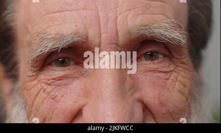 Extreme close-up macro portrait of wrinkled bearded face. Old senior beautiful man's eyes, smiling looking at camera. Brown eyes of elderly guy male model. Mature grandfather opening wide his eyes Stock Photo