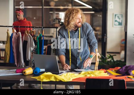 Smiling young caucasian fashion designer looking away while working at desk in creative office Stock Photo