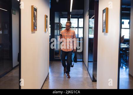 Caucasian mature businessman using smart phone while walking in corridor of creative office Stock Photo
