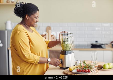 Side view portrait of young black woman using blender while making healthy smoothie at home kitchen, copy space Stock Photo