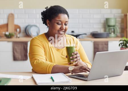 Portrait of smiling black woman drinking smoothie and using laptop at home Stock Photo