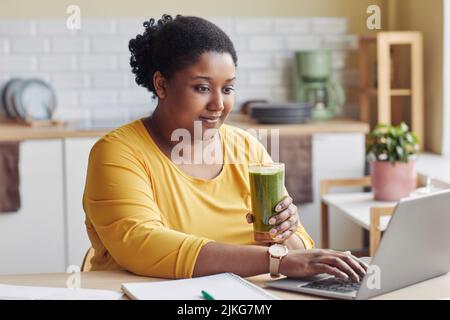 Portrait of overweight black woman drinking smoothie and using laptop at home Stock Photo