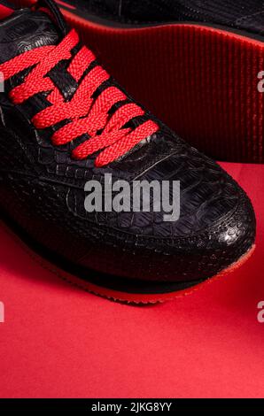 Fashionable snakeskin leather sneakers. Black sneakers with bright red bootlaces against a black and red background. Stock Photo