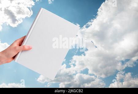 Hand with book mockup on blue cloudy sky background. Bible, inspiring literature concept. High quality photo Stock Photo