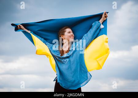 Happy free ukrainian woman with national flag on dramatic sky background. Portrait of lady in blue embroidery vyshyvanka shirt. Ukraine, independence Stock Photo