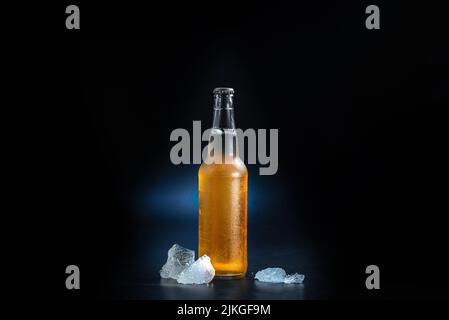 A bottle of beer with condensation droplets on a black and blue background. Cold beer bottle with pieces of ice near the beer bottle. A bottle of beer Stock Photo