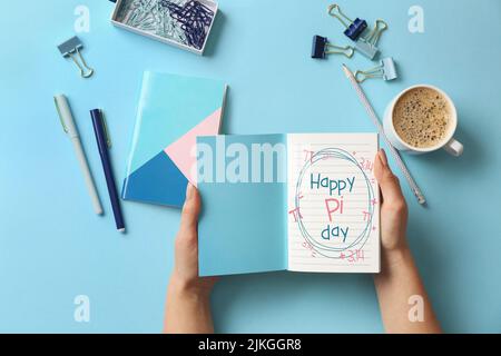 Female hands holding notebook with text HAPPY PI DAY, cup of coffee and stationery on light blue background Stock Photo