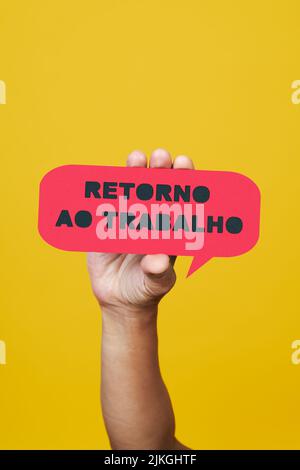 closeup of a man holding a red paper speech bubble with the text back to work written in portuguese, on a yellow background Stock Photo