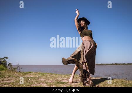 young latin caucasian tourist woman, wearing a long dress and black hat, is dancing on the edge of a cornice on the banks of the rio de la plata in ar Stock Photo