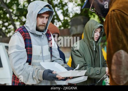 Homeless man holding plastic plate while volunteer putting food on it, he visiting charity community for homeless people Stock Photo