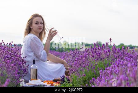 a girl holds a glass of white wine against the backdrop of a lavender field. A girl drinks wine in a lavender field. selective focus Stock Photo