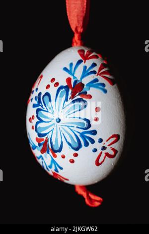 A vertical closeup of a handmade Easter egg hanging on a red ribbon against a black background Stock Photo
