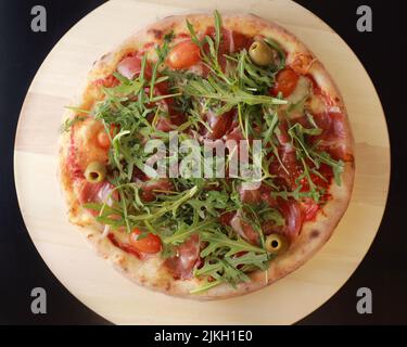 An appetizing pizza with ham, green olive and tomatoes dressed with the arugula on a wooden tray Stock Photo