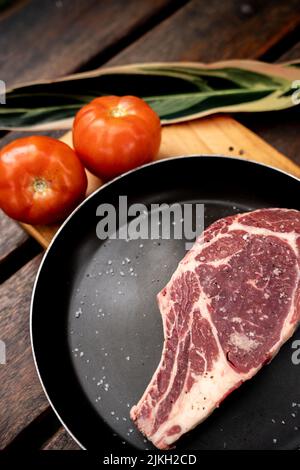 A vertical shot of a slice of raw red steak on a black pan on a board with tomatoes Stock Photo