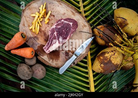 A top view of a raw steak on a wooden log board with a knife, french fries, carrots, and potatoes on a palm leaf background Stock Photo