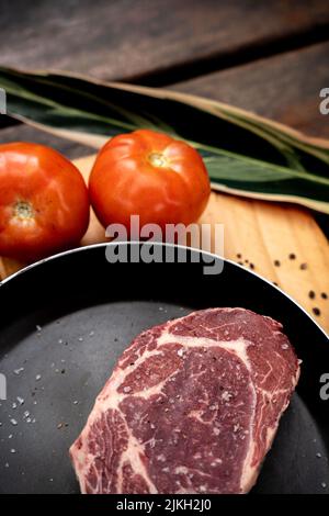 A vertical shot of a slice of raw red steak on a black pan on a board with tomatoes Stock Photo