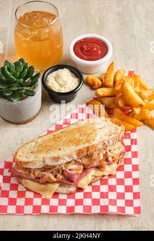 A vertical closeup of a sandwich with ham and sauerkraut on a checkered napkin next to fried potato, sauces, and orange juice Stock Photo